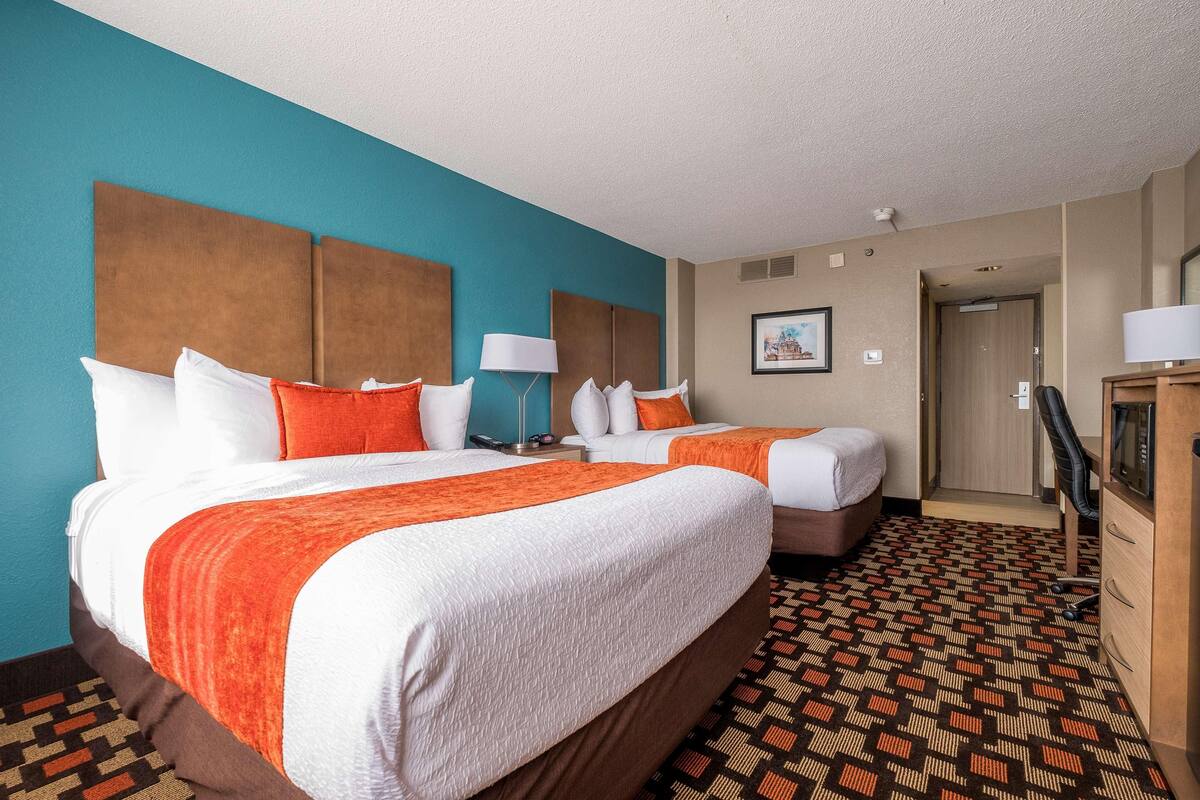 A hotel room with two beds, a patterned carpet, a wooden wardrobe, and a door leading to another room.