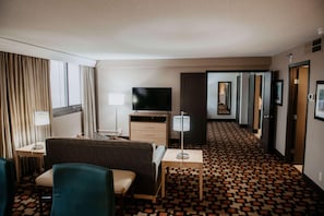 A hotel room with a sofa, a TV, a lamp, a patterned carpet, and a hallway leading to other rooms.