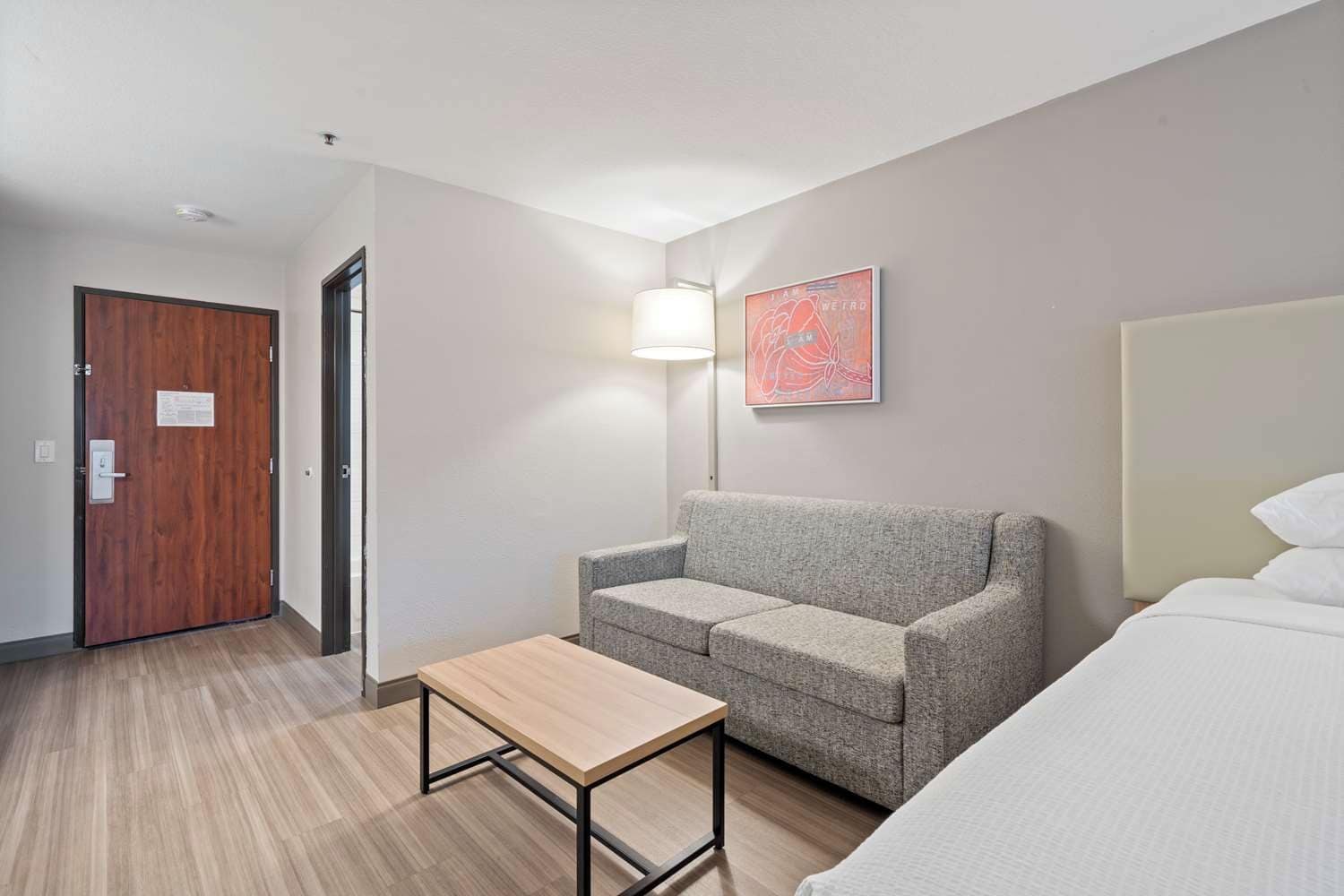 A hotel room with a wooden door, a grey sofa, a small table, a lamp, and a bed.