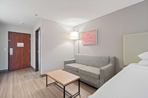 Una camera d'albergo con una porta in legno, un divano grigio, un tavolino, una lampada e un letto.