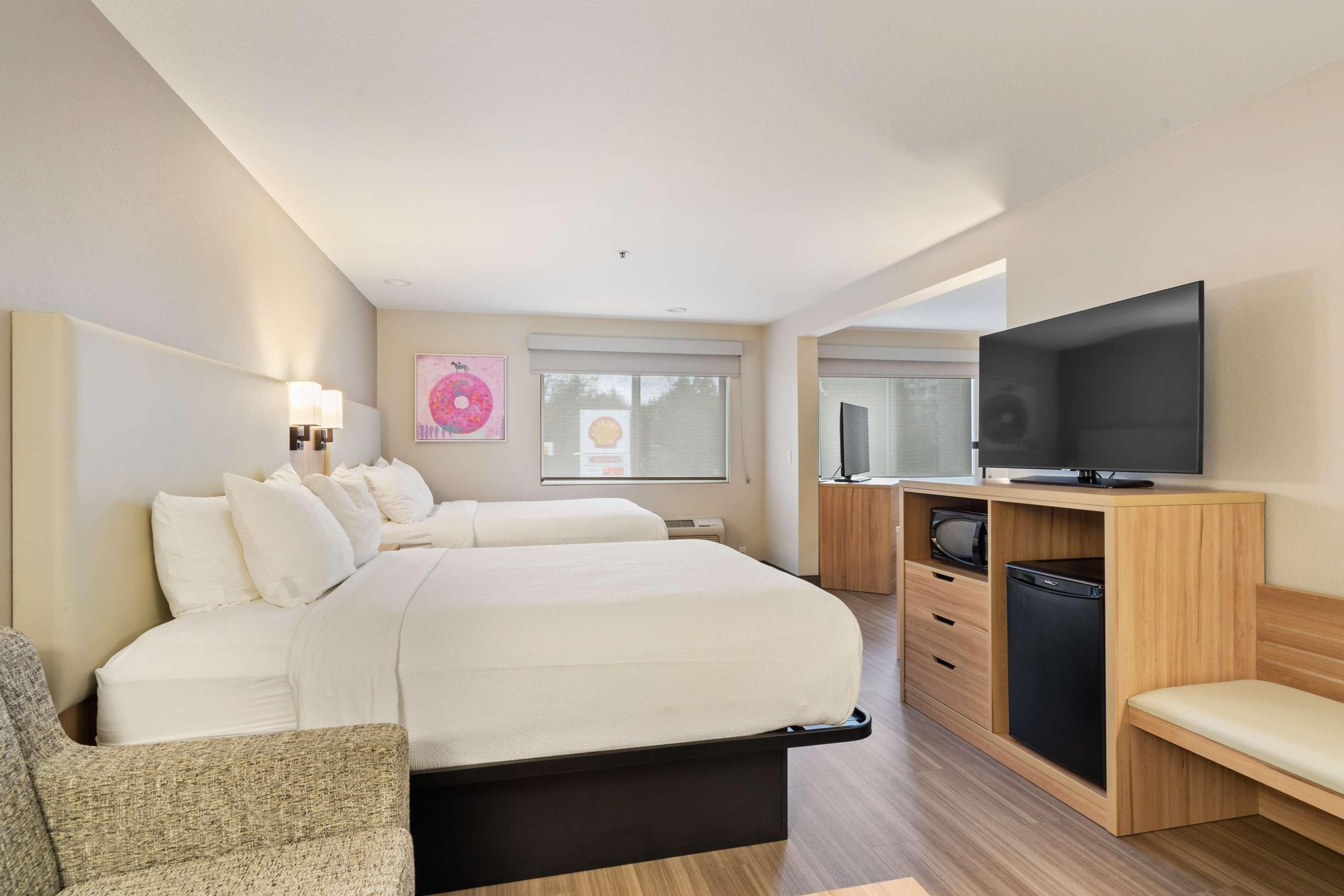 A hotel room with a large bed, a TV, a desk with a computer, and a window with blinds.