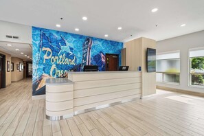 Un'area reception di un hotel dal design moderno, con una scrivania a forma di curva, un grande murale con la scritta 'Portland One' e vista all'esterno attraverso ampie finestre.