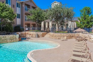 A pool area with lounge chairs, a stone wall, and a building with balconies in the background. - Lubbock West Inn and Suites (Lubbock)