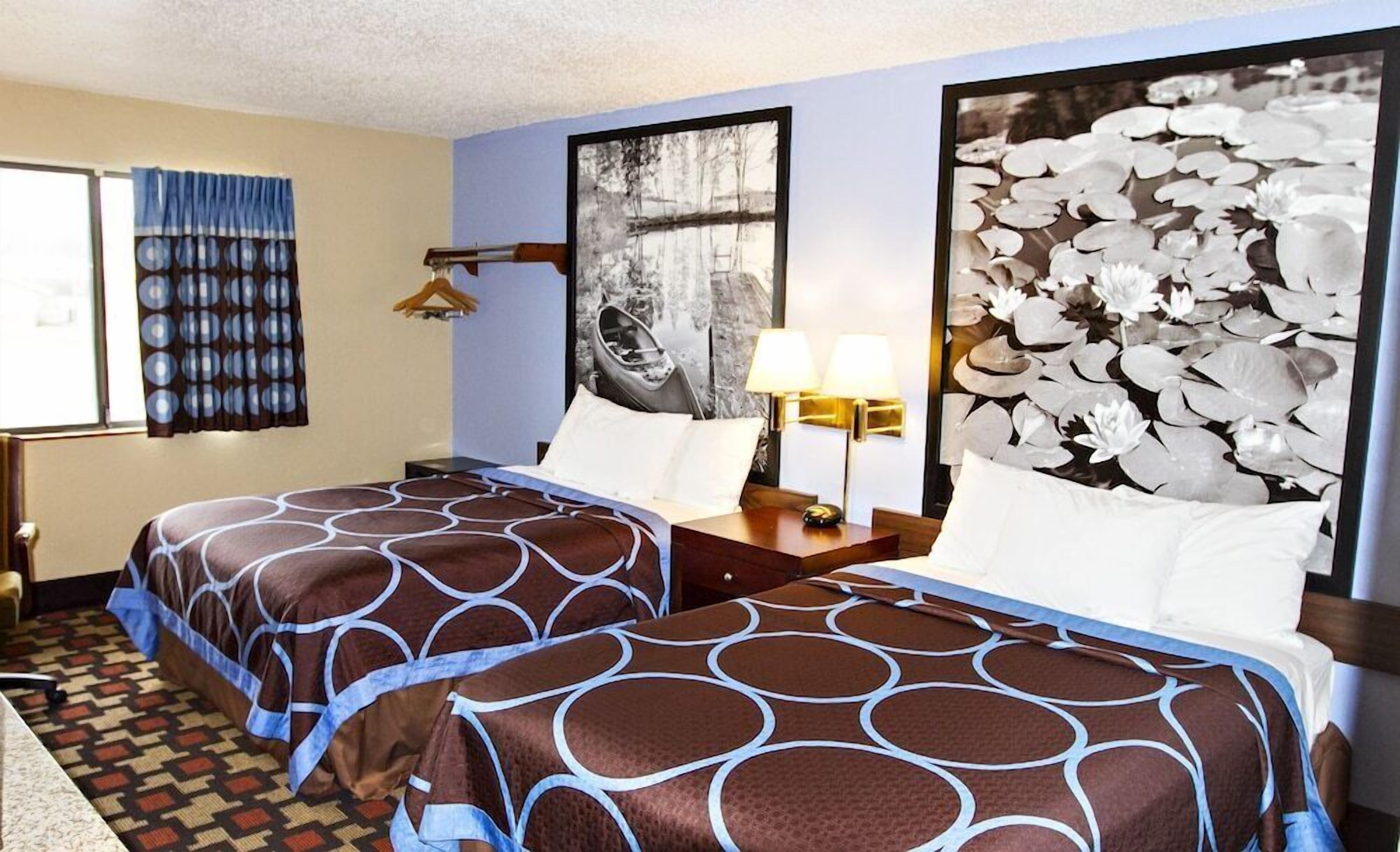 A hotel room with two beds, a window with curtains, a lamp, and a large abstract painting on the wall.
