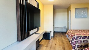 Een kamer met een wandgemonteerde flatscreen-tv, een bed met een decoratieve spreideken, een kleine verwarming en een luchtverkoeleenheid op de muur.