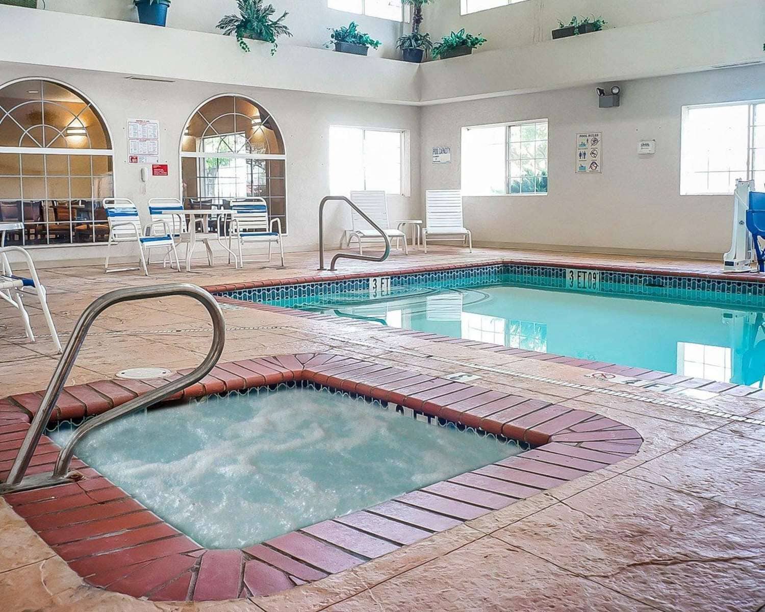An indoor swimming pool with a hot tub, lounge chairs, and a sunlit room with arched windows.