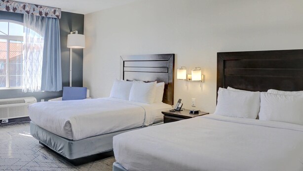 A hotel room with two beds, a bedside table with a lamp, a wall-mounted phone, and a window with curtains. - Quality Inn & Suites Roswell (Roswell)
