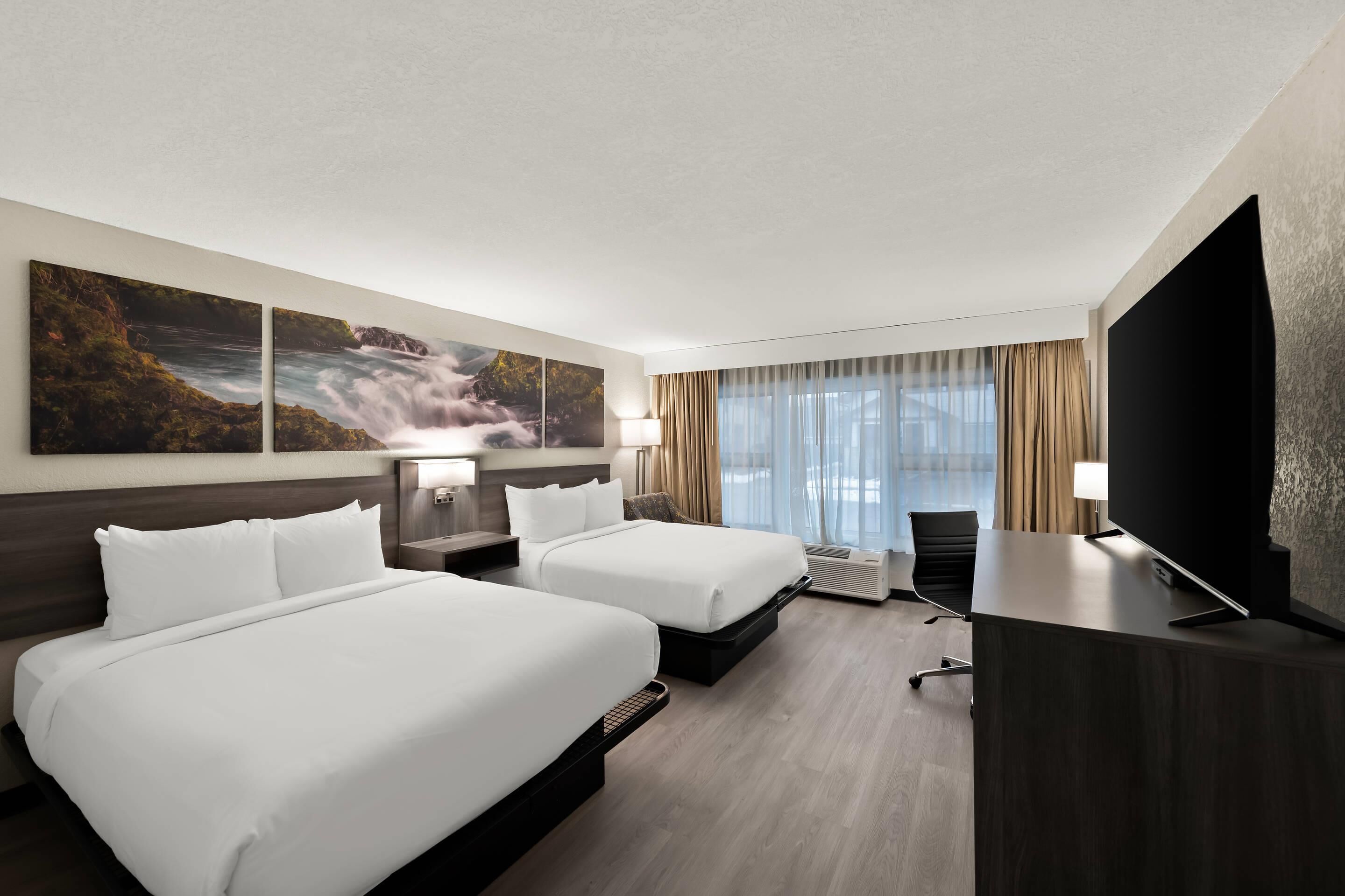 A hotel room with two beds, a desk, a TV, and a large window with curtains.