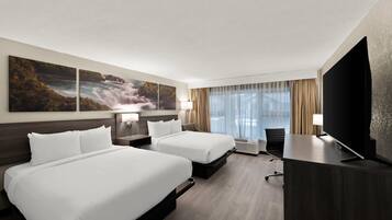 A hotel room with two beds, a desk, a TV, and a large window with curtains.