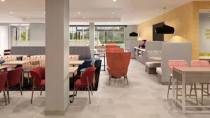 A modern dining area with tables and chairs, a central seating area with a sofa, and a wall-mounted light fixture. - Home2 Suites by Hilton Somerset (Somerset)