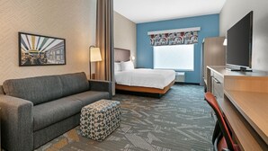 A hotel room with a bed, a sofa, a desk, a television, and a patterned carpet. - Home2 Suites by Hilton Somerset (Somerset)