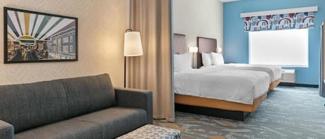 A hotel room with a sofa, a bed with two pillows, a patterned ottoman, a wall-mounted lamp, and a cityscape painting.