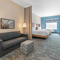 Habitación de hotel con sofá, cama con dos almohadas, otomana estampada, lámpara de pared y pintura de un paisaje urbano.