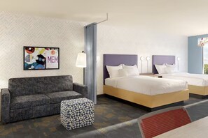 A hotel room with a grey sofa, a patterned ottoman, a bed with white bedding, and a wall-mounted artwork. - Home2 Suites by Hilton Somerset (Somerset)