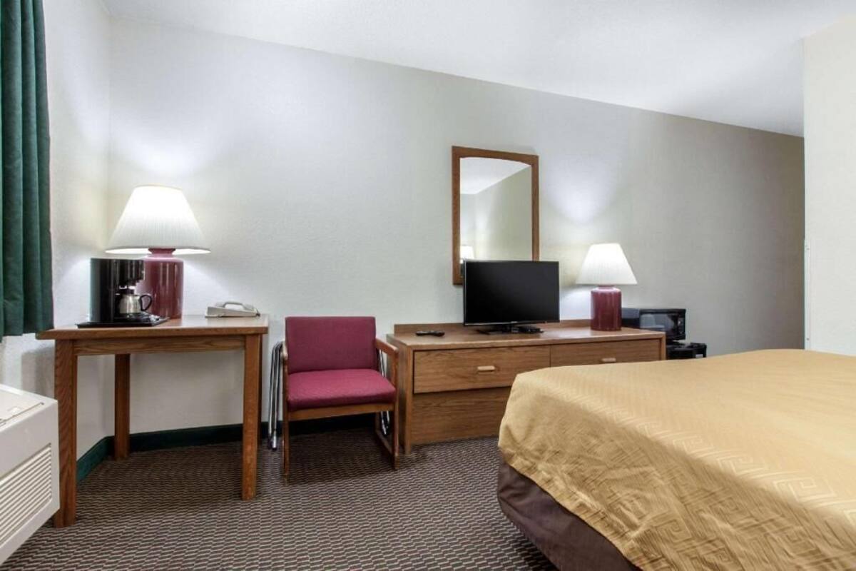 A hotel room with a bed, a desk with a lamp, a chair, a television, and a dresser.