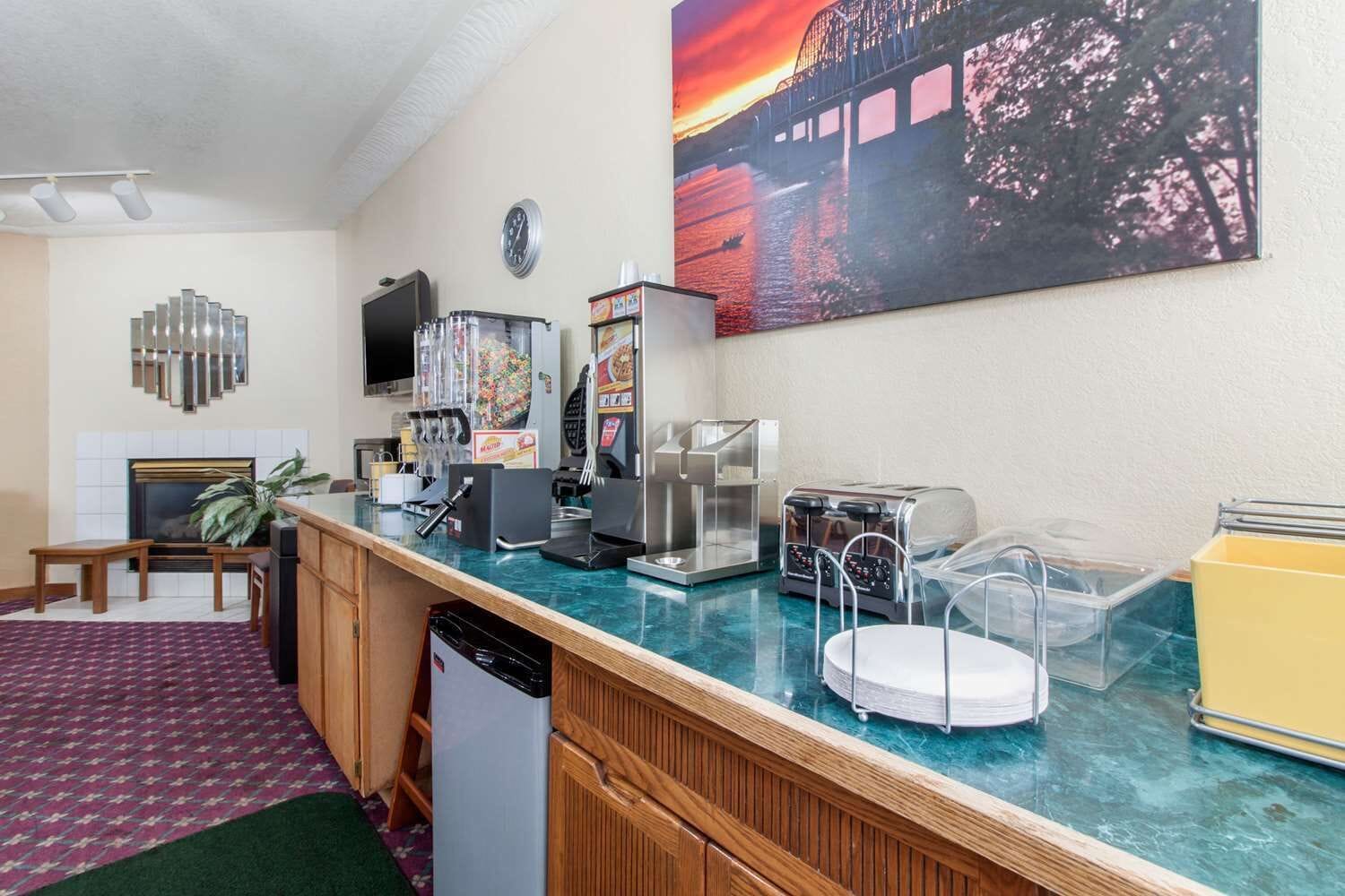A hotel room with a kitchenette, a flat-screen TV, a painting of a sunset, and a green carpet.