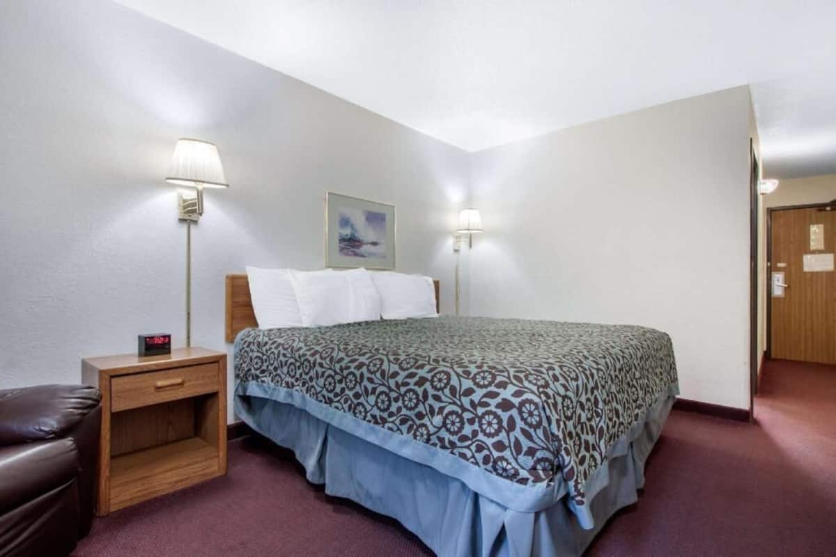 A hotel room with a bed, two bedside lamps, a nightstand, a clock, a framed picture on the wall, and a door.