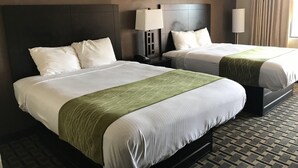 Duas camas de casal com roupa de cama branca e detalhes verdes, uma mesa de cabeceira com abajur, e uma janela com cortinas.
