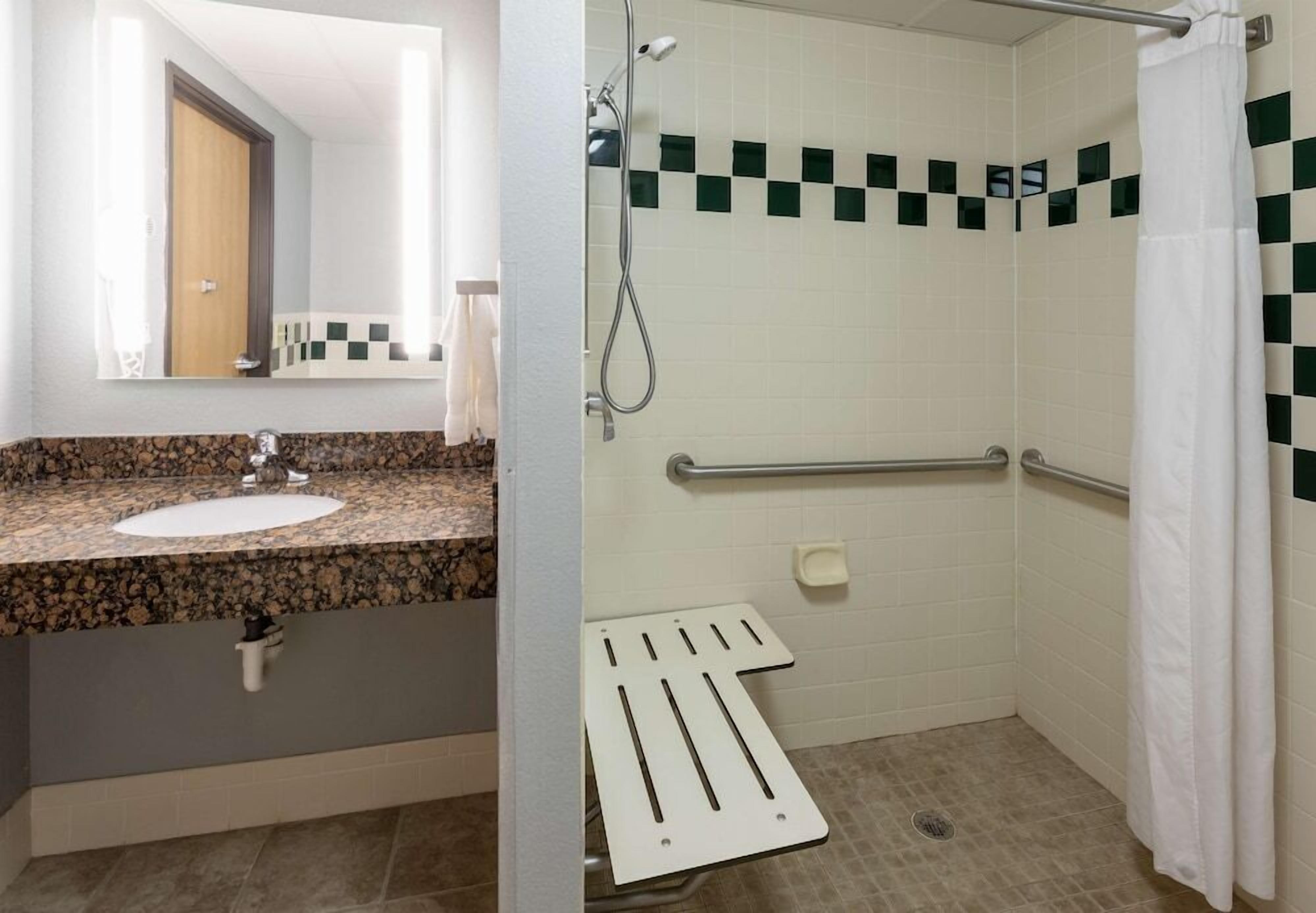 A bathroom with a granite countertop sink, a mirror, and a shower with a handrail.