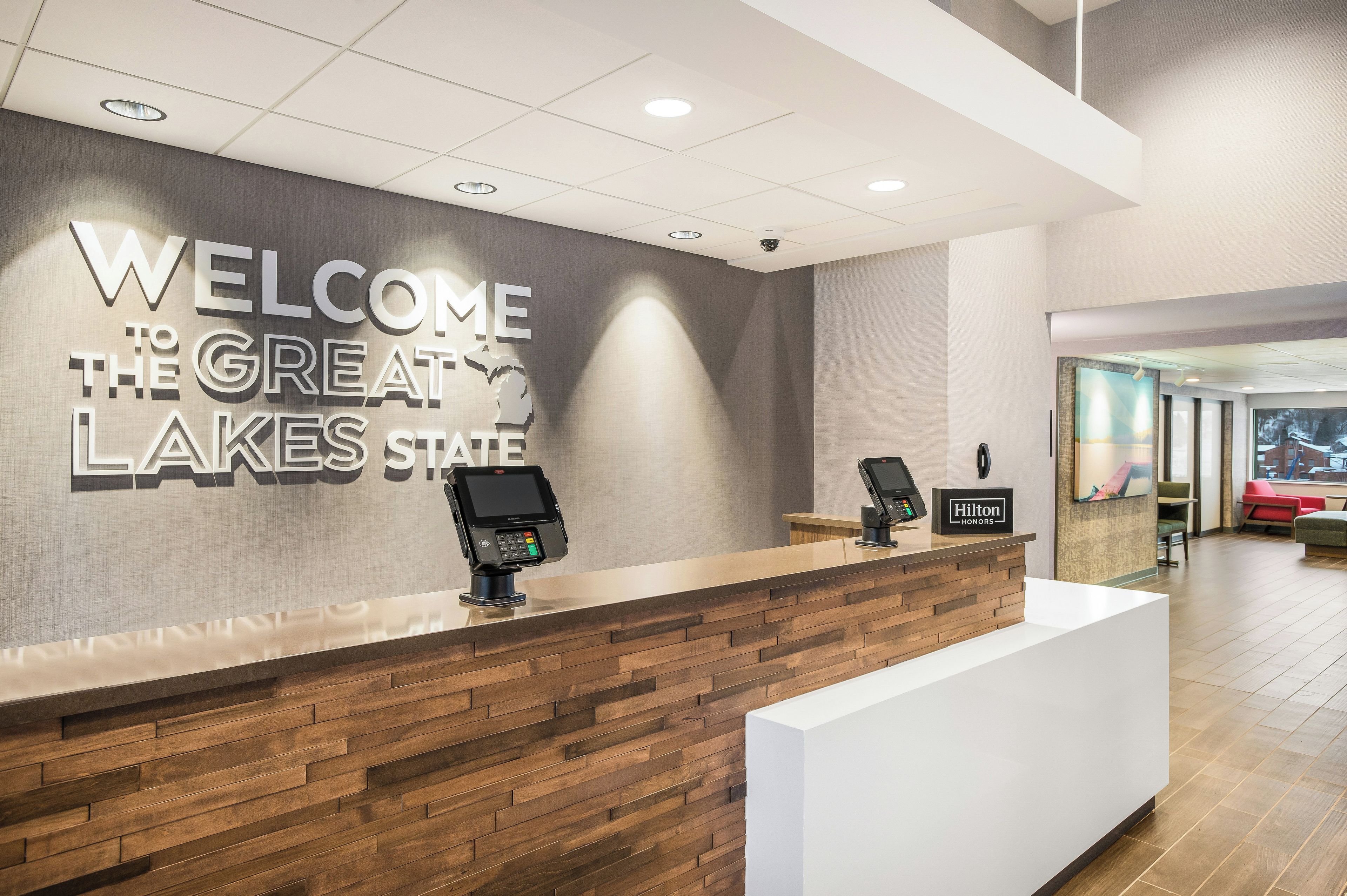 Un mostrador de recepción con un cartel que dice 'Welcome to the Great Lakes State', un quiosco digital y una placa de 'Hilton Honors'.