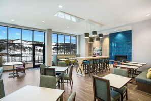 A modern hotel lobby with a large window offering a view of a snowy landscape, a seating area with tables and chairs, and a blue-tiled wall. - Hampton Inn & Suites Houghton (Houghton)
