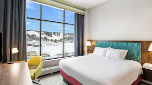A hotel room with a large bed, a desk, a chair, and a view of a snowy landscape through a large window. - Hampton Inn & Suites Houghton (Houghton)
