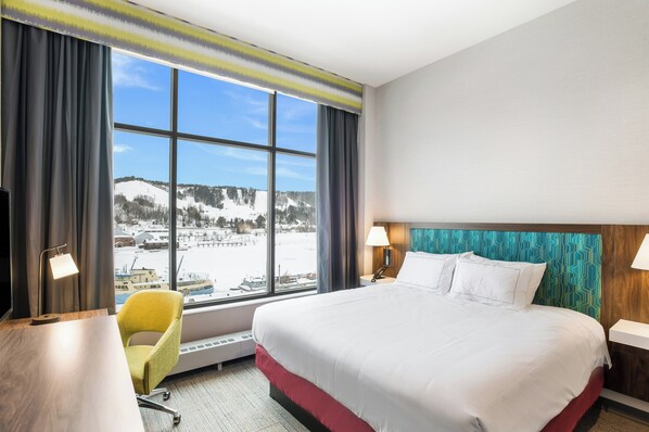 A hotel room with a large bed, a desk, a chair, and a view of a snowy landscape through a large window. - Hampton Inn & Suites Houghton (Houghton)