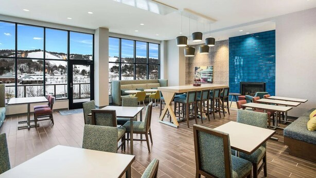 A modern dining area with large windows, a fireplace, and a variety of seating options. - Hampton Inn & Suites Houghton (Houghton)
