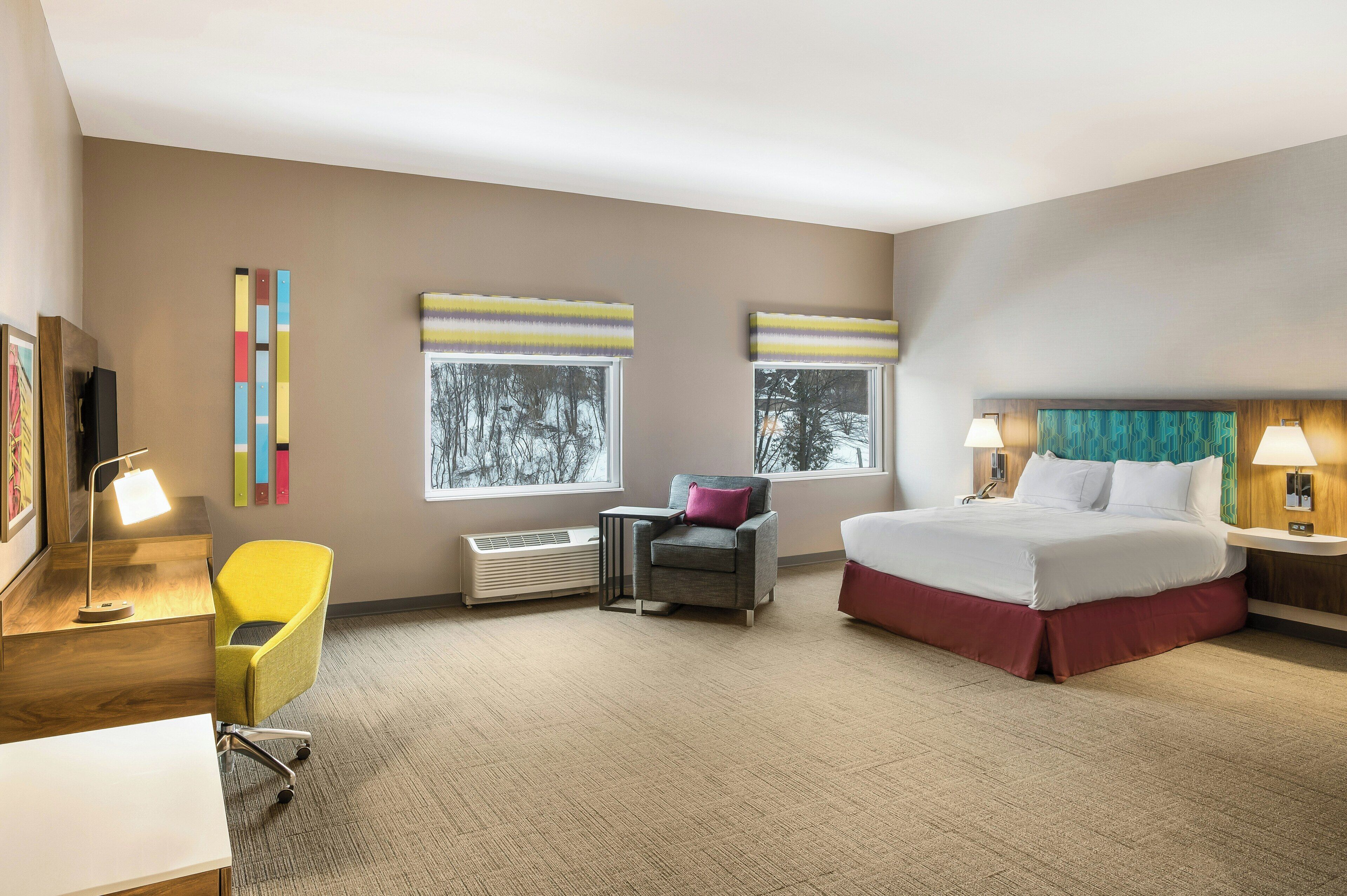 Habitación de hotel con cama, escritorio, silla, dos ventanas con persianas, calefactor y televisor montado en la pared.