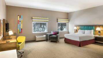 Habitación de hotel con cama, escritorio, silla, dos ventanas con persianas, calefactor y televisor montado en la pared.