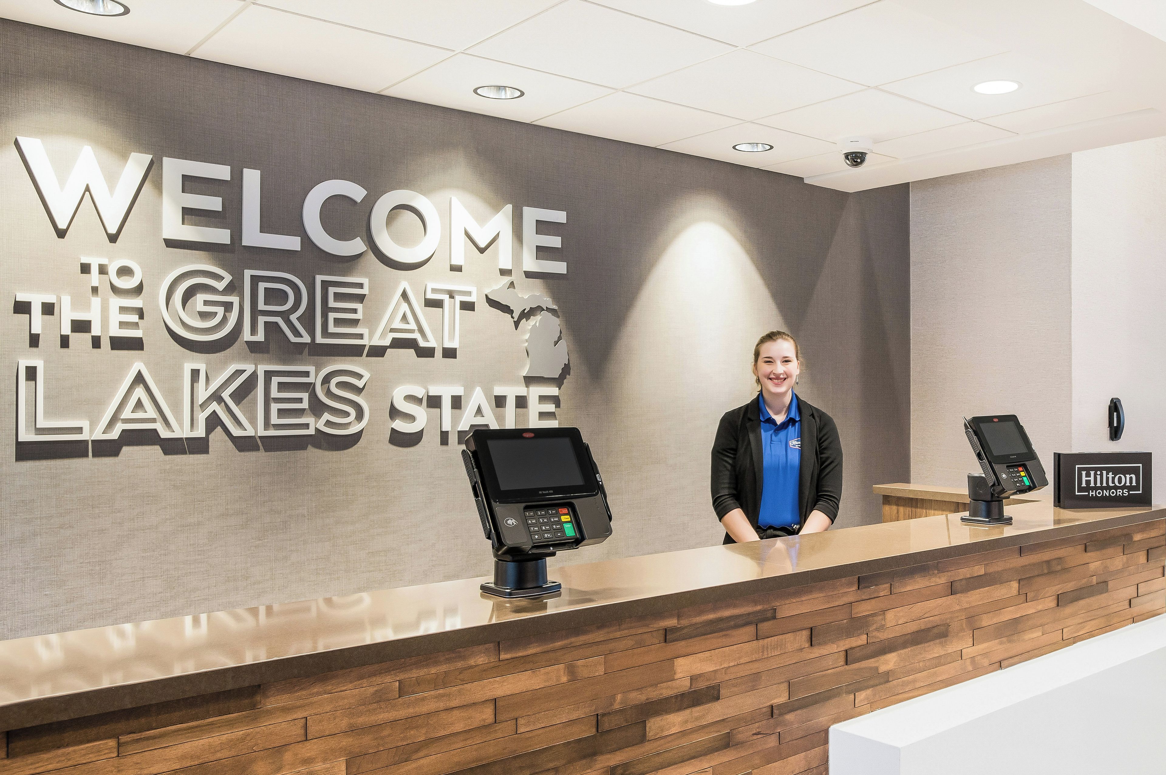 A reception desk with a 'WELCOME THE GREAT LAKES STATE' sign, a digital kiosk, and a 'Hilton Honors' sign.