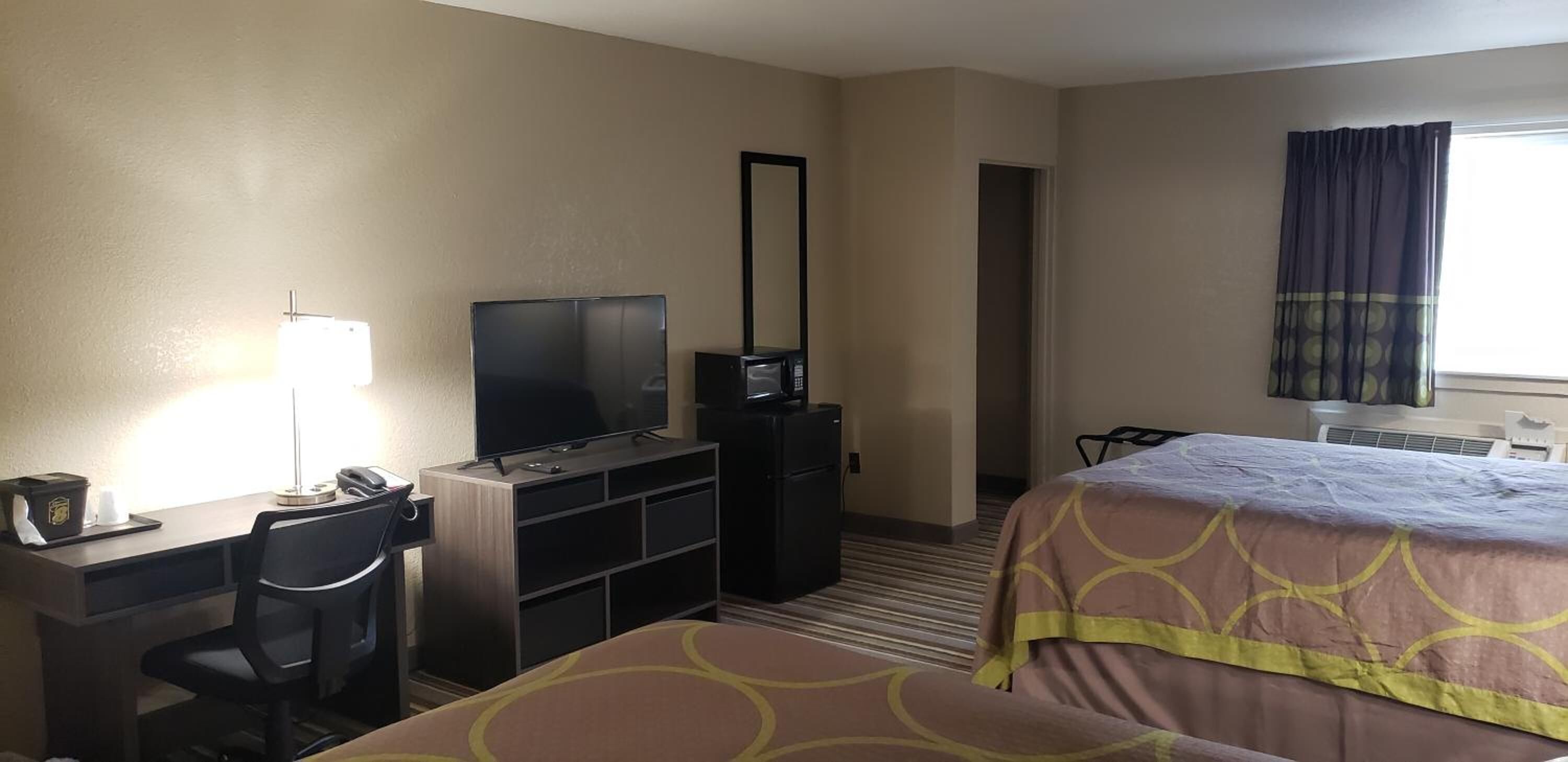 Een hotelkamer met een bureau, een televisie, een dressoir, een bed met een geborduurde spreideken en een raam met gordijnen.