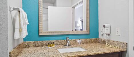 A bathroom with a granite countertop, a sink, a mirror, a towel rack, and a hairdryer.