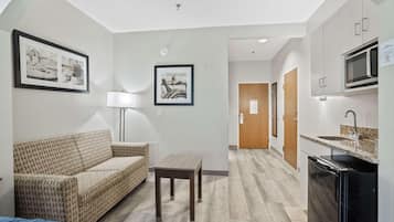 A compact hotel room with a sofa, a small table, a lamp, a kitchenette with a microwave, and framed pictures on the walls.