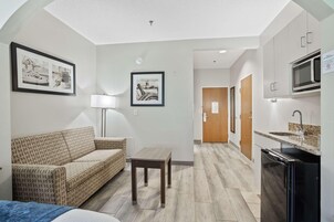 Habitación de hotel compacta con sofá, una mesita, lámpara, kitchenette con microondas y cuadros en las paredes.
