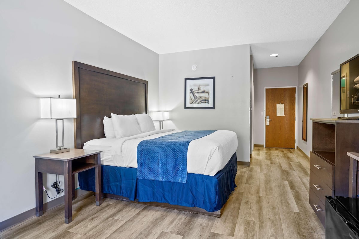A hotel room with a bed, bedside tables, a desk, a mirror, a framed picture on the wall, and a door leading to another room.