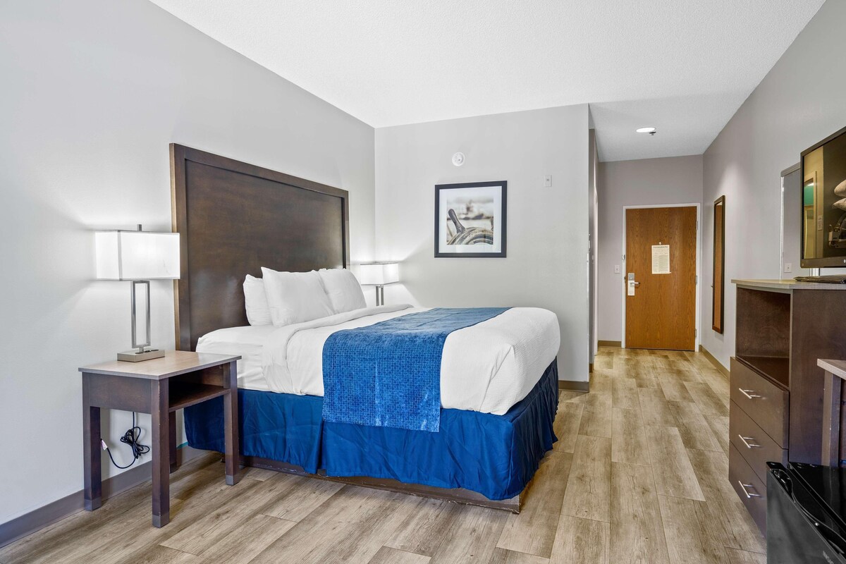 A hotel room with a bed, bedside tables, a desk, a mirror, a framed picture on the wall, and a door leading to another room.