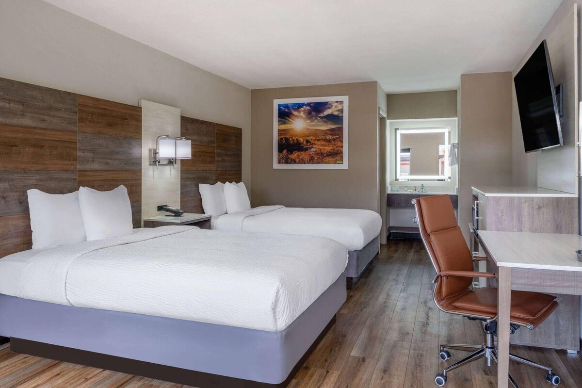 A hotel room with two beds, a desk, a chair, a TV, a painting, and a bathroom mirror.