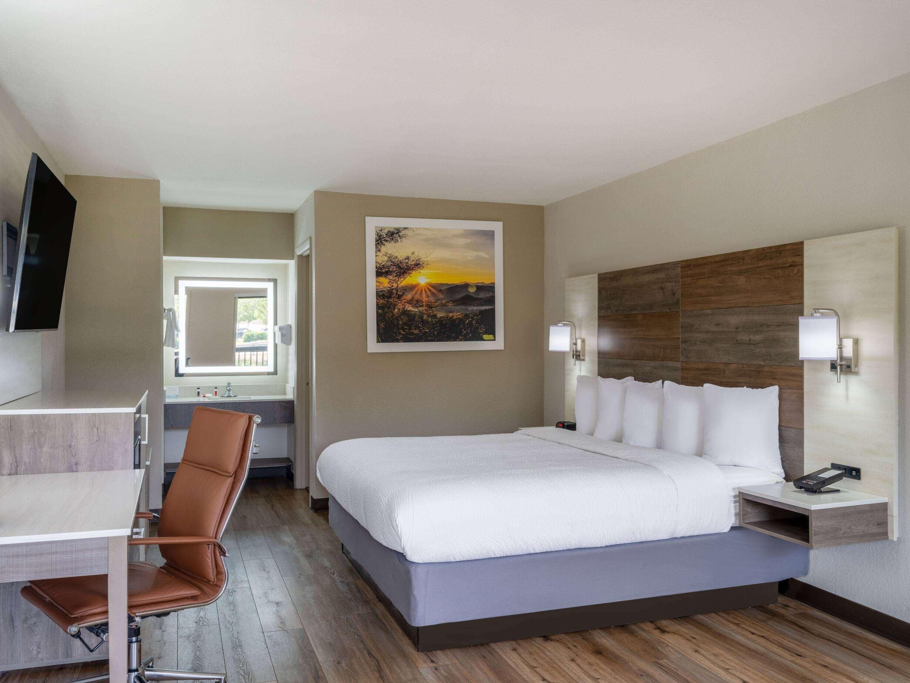 A hotel room with a bed, a desk, a chair, a TV, a painting, and a nightstand with a phone.