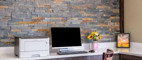 Un espacio de trabajo con una impresora, un monitor de computadora, un teclado, un florero con flores y un cuadro en un escritorio.