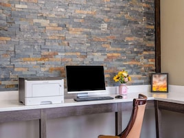 Un espacio de trabajo con una impresora, un monitor de computadora, un teclado, un florero con flores y un cuadro en un escritorio.