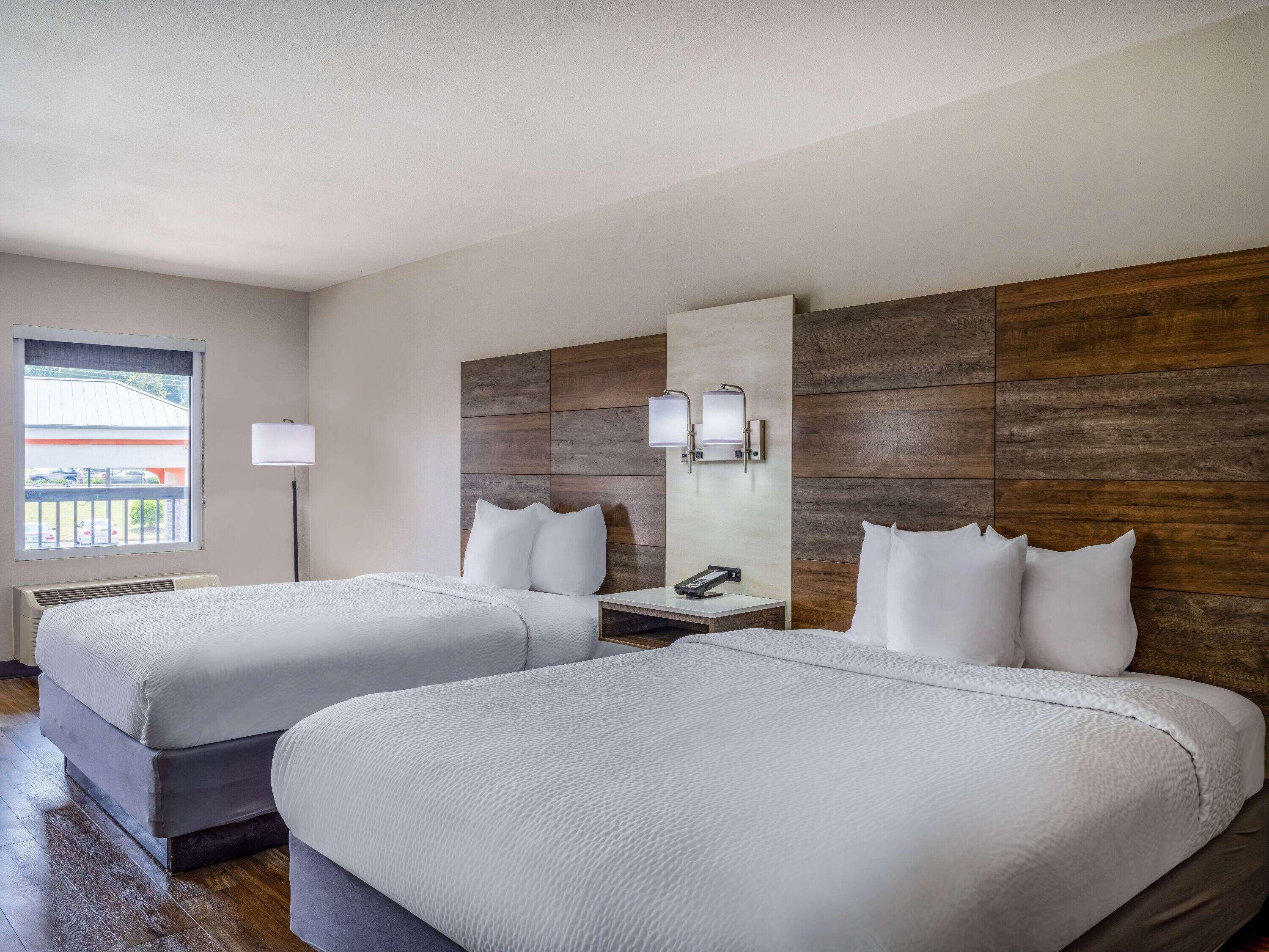 A hotel room with two beds, a nightstand, a lamp, and a window with a view.