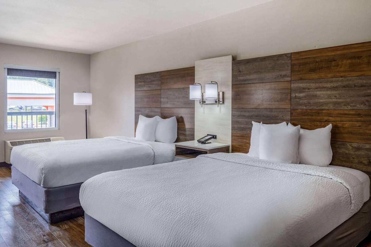 A hotel room with two beds, a nightstand, a lamp, and a window with a view.