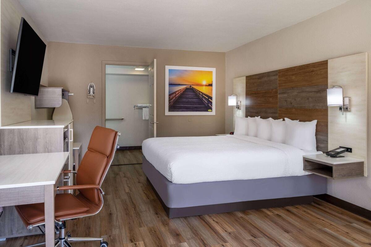 A hotel room with a bed, desk, chair, and a painting of a sunset over a pier.