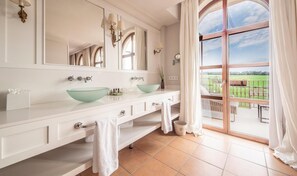 Baño con un espejo grande, dos lavamanos, una bañera y vista a un campo verde a través de una puerta de cristal.
