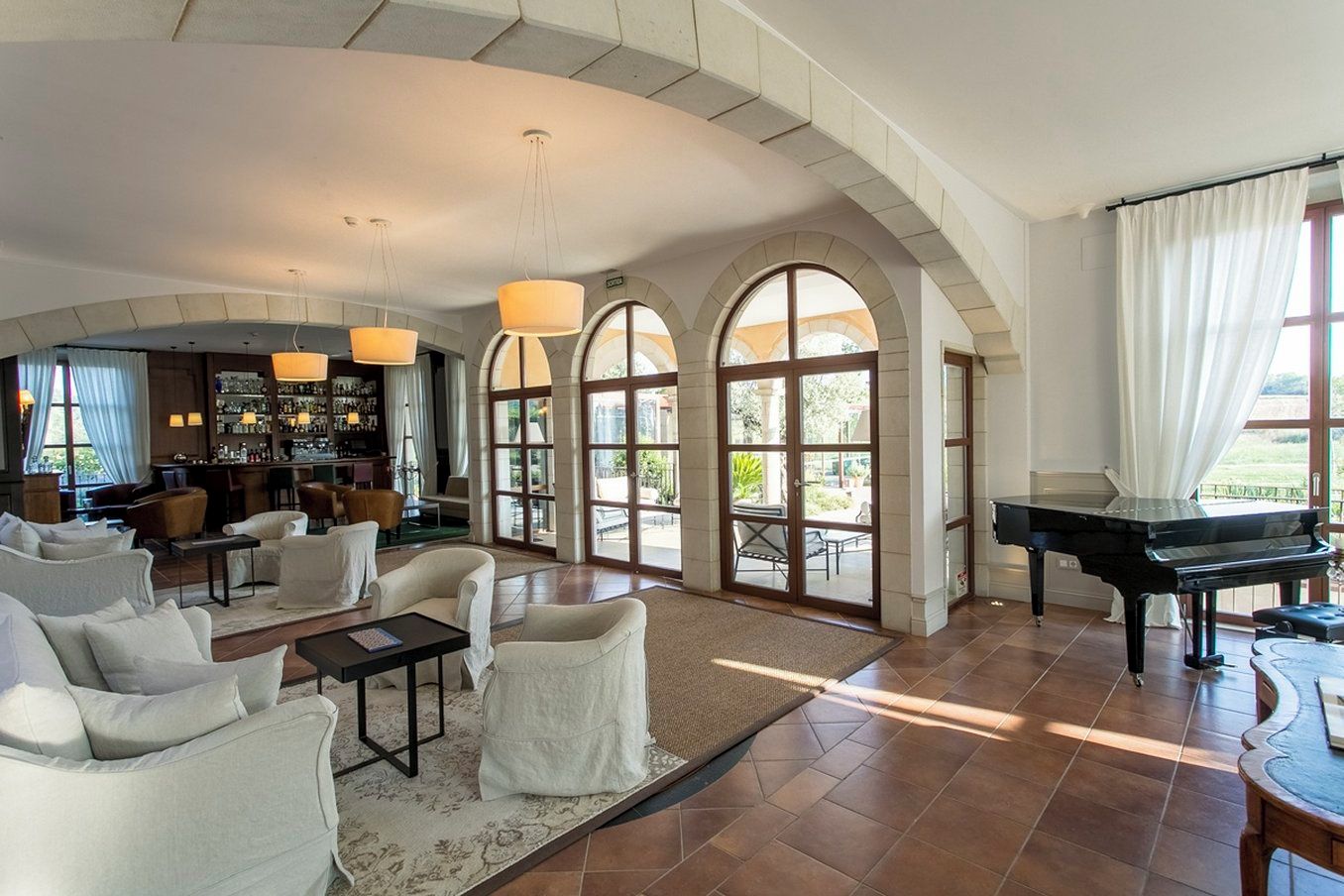 A spacious living room with a grand piano, comfortable seating, a bar area, and large arched windows.