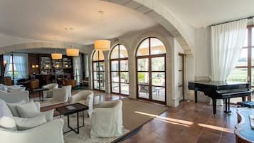 A spacious living room with a grand piano, comfortable seating, a bar area, and large arched windows.