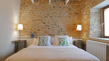 A bedroom with a stone wall, a bed with a beige duvet, two bedside tables with lamps, and a window with a view.