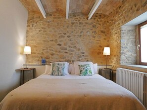Un dormitorio con pared de piedra, una cama con cubrecama color beige, dos mesitas de noche con lámparas y una ventana con vistas.