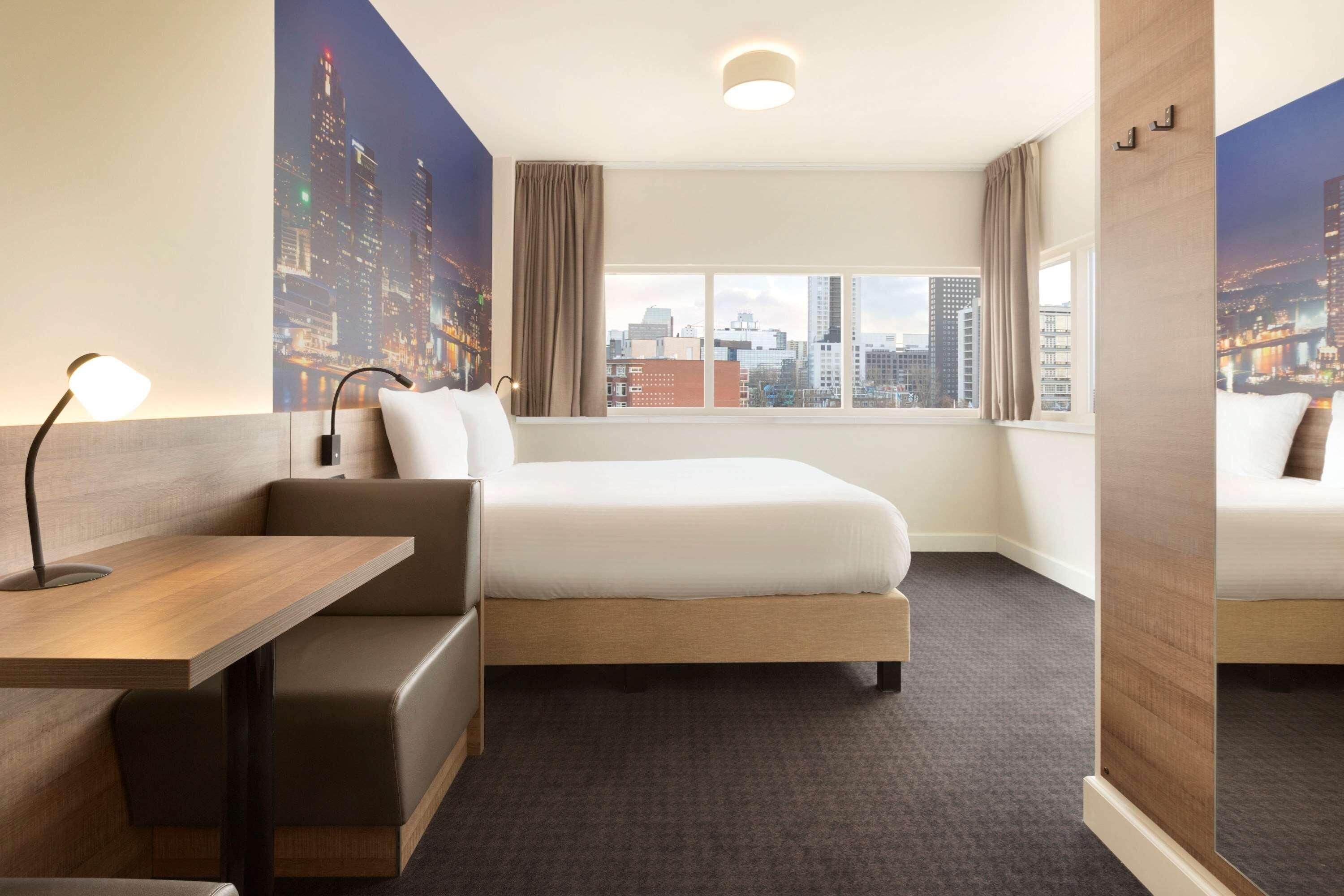 A hotel room with a bed, a desk, a chair, a window with a cityscape view, and a large poster on the wall.
